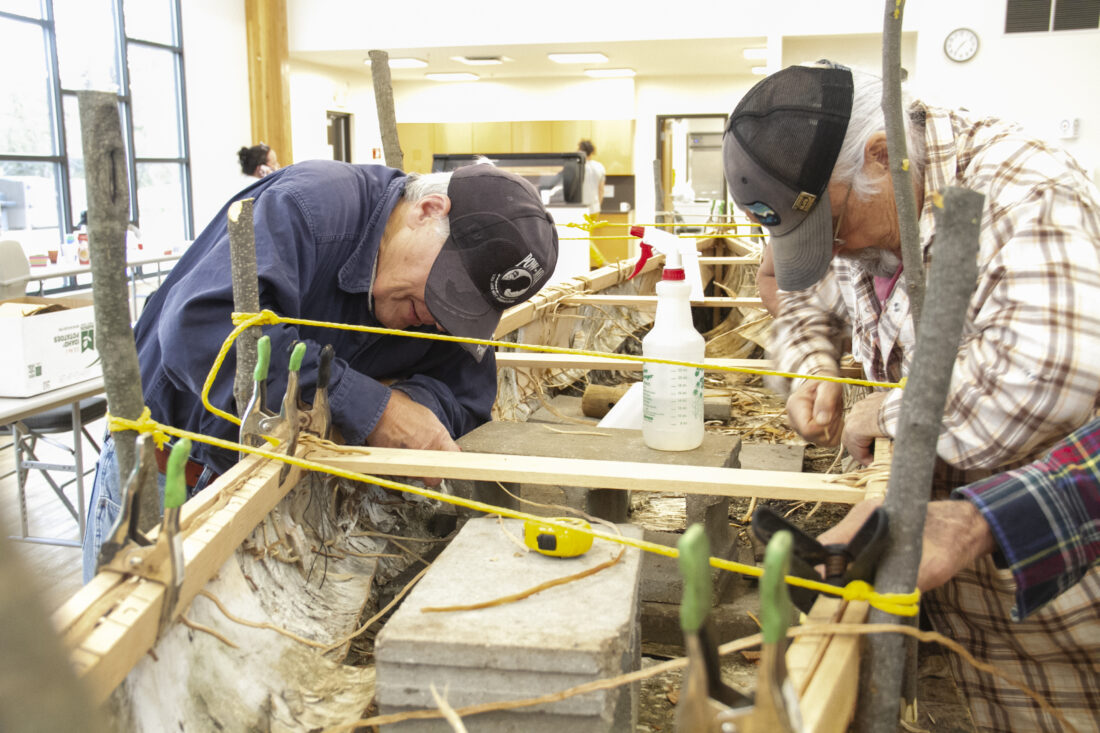 Ernie Demmon and Larry Godfrey of Gladstone lean over the gunnels of an in-process canoe as they work to pull through strips of spruce root that hold pieces together, like heavy-duty stitching. (R. R. Branstrom | Daily Press)